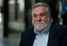 Older man with gray hair and beard in a navy blazer and light blue shirt, seated for an interview.