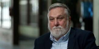 Older man with gray hair and beard in a navy blazer and light blue shirt, seated for an interview.