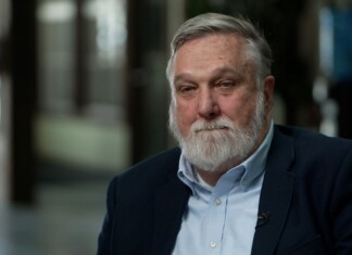 Older man with gray hair and beard in a navy blazer and light blue shirt, seated for an interview.