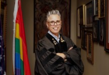 Smiling person in a black academic gown with pins, arms crossed, standing in a hallway lined with framed art and flags (U.S. flag and rainbow Pride flag).