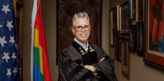 Smiling person in a black academic gown with pins, arms crossed, standing in a hallway lined with framed art and flags (U.S. flag and rainbow Pride flag).