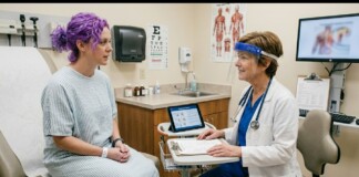 Patient with purple hair sits on a medical exam chair wearing a light striped gown while a doctor in blue scrubs and a face shield reviews notes at a mobile cart in a clinic examination room.