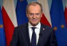 Smiling man in a navy suit giving a press-address, with EU and Polish flags in the background.