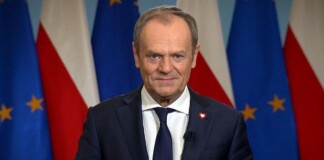 Smiling man in a navy suit giving a press-address, with EU and Polish flags in the background.
