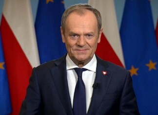Polish Prime Minister Apologizes To The LGBTQ Community For Rejections And Humiliation Smiling man in a navy suit giving a press-address, with EU and Polish flags in the background.