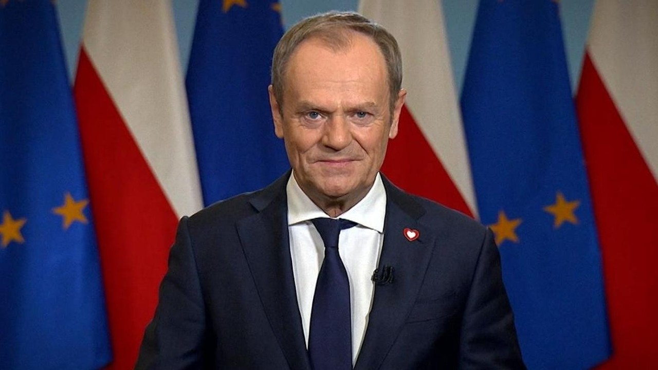 Smiling man in a navy suit giving a press-address, with EU and Polish flags in the background.