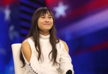 Smiling woman with long dark hair sits on a white chair on a stage with a blue‑red backdrop.
