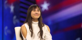 Smiling woman with long dark hair sits on a white chair on a stage with a blue‑red backdrop.