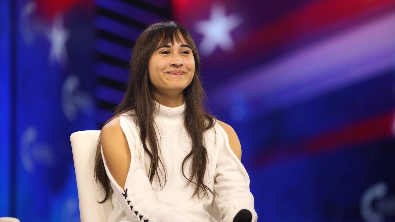 University of Washington Postpones Turning Point USA Event After The Trans Student Killing Smiling woman with long dark hair sits on a white chair on a stage with a blue‑red backdrop.