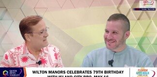 Two men sit side by side and smile during a TV talk show in a studio with a geometric pastel backdrop; a caption banner at the bottom mentions Wilton Manors' birthday event.