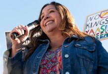 Woman speaking into a handheld microphone at an outdoor rally, smiling, wearing a denim jacket and colorful top with a protest sign visible in the background.