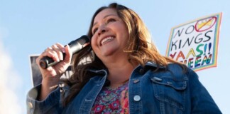 Woman speaking into a handheld microphone at an outdoor rally, smiling, wearing a denim jacket and colorful top with a protest sign visible in the background.