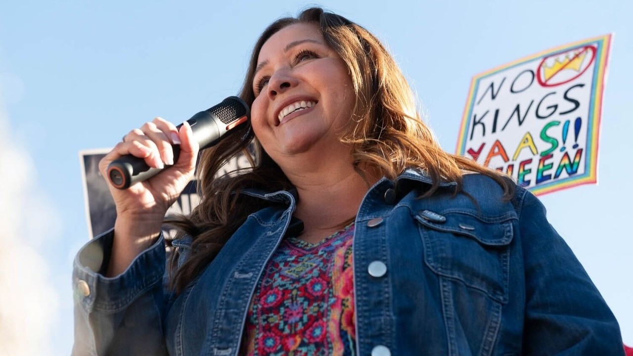 Woman speaking into a handheld microphone at an outdoor rally, smiling, wearing a denim jacket and colorful top with a protest sign visible in the background.