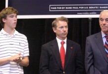 Three men stand on a stage: a young man in a white striped polo on the left, a suited man with a red tie in the middle, and an older man in a gray pinstripe suit on the right, with a campaign banner and American flag in the background.