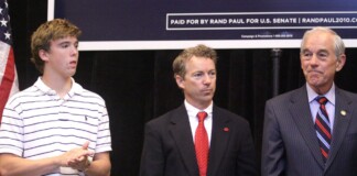 Three men stand on a stage: a young man in a white striped polo on the left, a suited man with a red tie in the middle, and an older man in a gray pinstripe suit on the right, with a campaign banner and American flag in the background.
