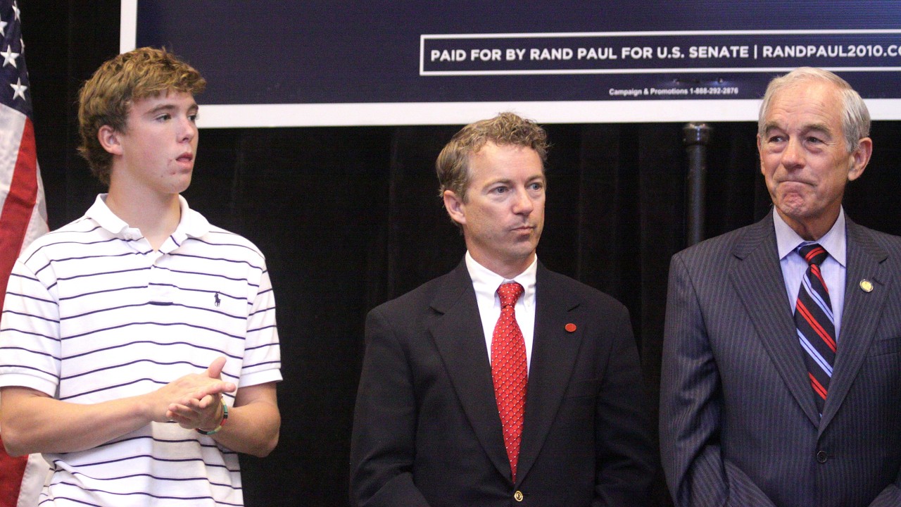 Three men stand on a stage: a young man in a white striped polo on the left, a suited man with a red tie in the middle, and an older man in a gray pinstripe suit on the right, with a campaign banner and American flag in the background.