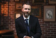 Man in a dark suit with a blue tie sits at a wooden desk in a brick-walled study, looking at the camera with a slight smile.