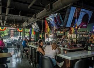 Bar interior with rainbow flags hanging by windows, TVs overhead, and patrons seated along a long white bar with bottles behind it.