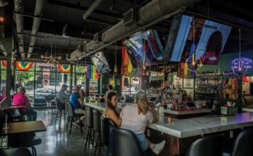 Nashville LGBTQ Bar Targeted Twice in Less Than 48 Hours Ahead of Pride Month Bar interior with rainbow flags hanging by windows, TVs overhead, and patrons seated along a long white bar with bottles behind it.