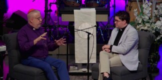 Two men in conversation on a stage, one in a purple shirt with an orange yarmulke and the other in a light blazer, seated on gray chairs with a microphone between them.