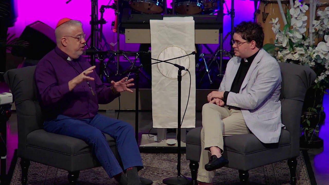 Two men in conversation on a stage, one in a purple shirt with an orange yarmulke and the other in a light blazer, seated on gray chairs with a microphone between them.