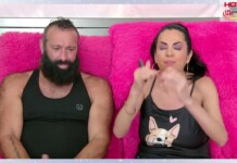 Man with a beard in a black tank top sits beside a woman with purple makeup on bright pink fuzzy chairs during a talk show.