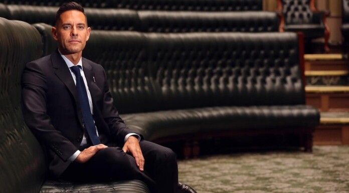Australian Tribunal Orders Parliament Member To Pay $100k For Vilifying Gay MP Man in a dark suit and blue tie seated on a tufted green leather bench in a formal room, looking at the camera.