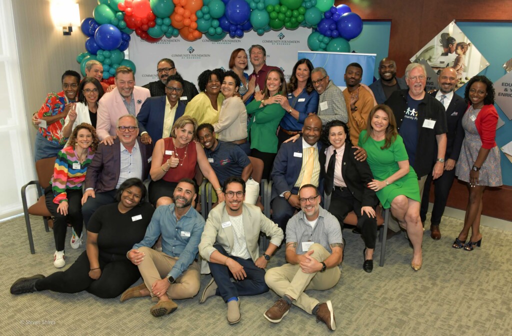 Diverse group of people posing for a group photo at an indoor event, colorful balloon arch behind them and many wearing name tags and smiles.