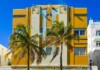Art Deco building with yellow and blue accents, tiled decorative panels, and palm trees against a bright blue sky.