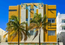 Art Deco building with yellow and blue accents, tiled decorative panels, and palm trees against a bright blue sky.