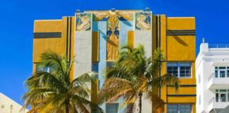 Art Deco building with yellow and blue accents, tiled decorative panels, and palm trees against a bright blue sky.