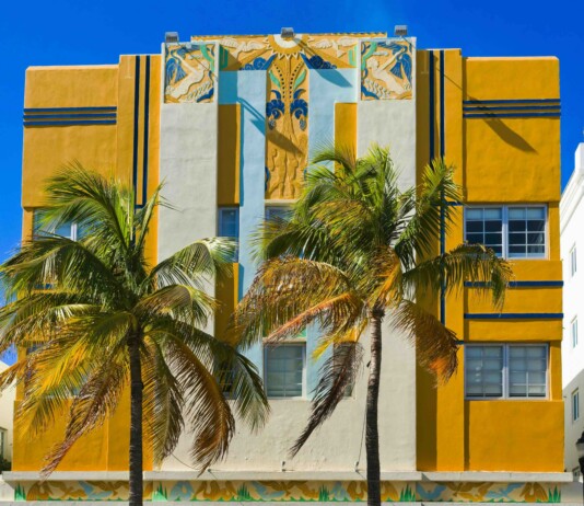 10 Iconic Location in Florida to Visit Art Deco building with yellow and blue accents, tiled decorative panels, and palm trees against a bright blue sky.
