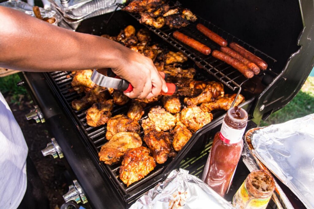 Hand using tongs to grill chicken wings and sausages on a barbecue, sauce bottle nearby.