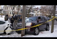 Winter residential crash scene: a maroon SUV and a white car damaged, police and responders behind yellow caution tape, snow on the ground.