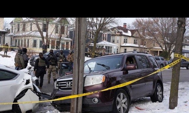 Winter residential crash scene: a maroon SUV and a white car damaged, police and responders behind yellow caution tape, snow on the ground.