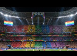 Fans in a stadium hold rainbow cards, creating a Pride banner across the stands beneath a large 'PRIDE' sign.