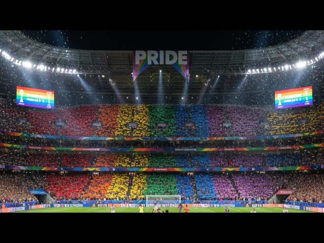 Fans in a stadium hold rainbow cards, creating a Pride banner across the stands beneath a large 'PRIDE' sign.