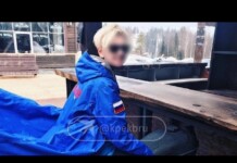 Person in a bright blue winter jacket with a Russian flag patch sits at a wooden railing outdoors in a snowy setting; face blurred. Readable watermark at center.
