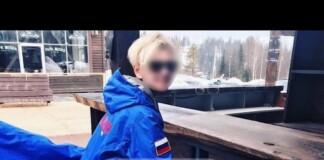 Person in a bright blue winter jacket with a Russian flag patch sits at a wooden railing outdoors in a snowy setting; face blurred. Readable watermark at center.
