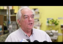 Older man with white hair speaks to reporters in a yellow-walled room, microphones in front of him.