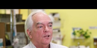 Older man with white hair speaks to reporters in a yellow-walled room, microphones in front of him.