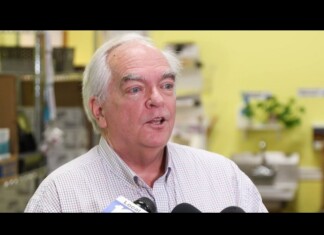 Older man with white hair speaks to reporters in a yellow-walled room, microphones in front of him.