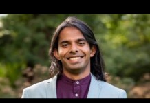 Smiling man with long dark hair in a light blue blazer and maroon shirt, outdoors in greenery