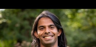 Smiling man with long dark hair in a light blue blazer and maroon shirt, outdoors in greenery