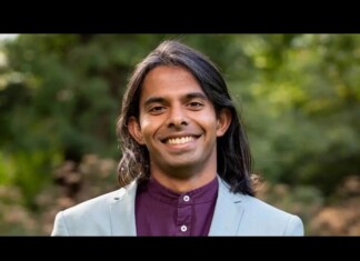 Smiling man with long dark hair in a light blue blazer and maroon shirt, outdoors in greenery