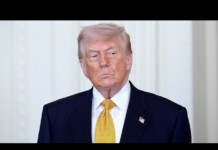 Former U.S. president Donald Trump standing in a dark suit and yellow tie, with a small American flag pin on the lapel.