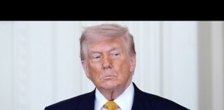 Former U.S. president Donald Trump standing in a dark suit and yellow tie, with a small American flag pin on the lapel.