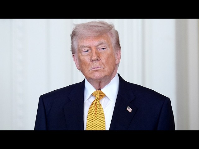 Former U.S. president Donald Trump standing in a dark suit and yellow tie, with a small American flag pin on the lapel.