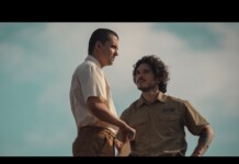 Two men in an outdoor setting stand close, one in a light shirt and the other in a tan uniform, seeming to have a serious conversation.