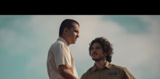 Two men in an outdoor setting stand close, one in a light shirt and the other in a tan uniform, seeming to have a serious conversation.